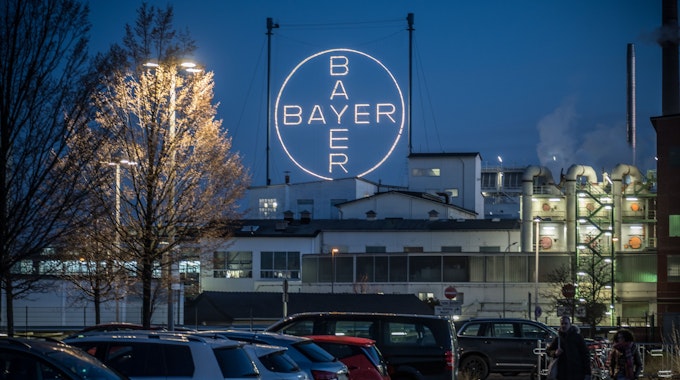 Das leuchtende Bayer-Kreuz ist aus einiger Entfernung vor parkenden Autos im Chempark Leverkusen abgebildet. Schornsteine qualmen im rechten Teil des Bildes, links befinden sich zwei Bäume.&nbsp;