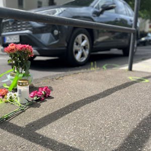 Eine Grabkerze und Rosen stehen am Freitag, 13.05.2022, am Unfallort auf der Aachener Straße in Höhe der Haltestelle Melaten, wo tags zuvor eine 43-jährige Fußgängerin überfahren wurde.