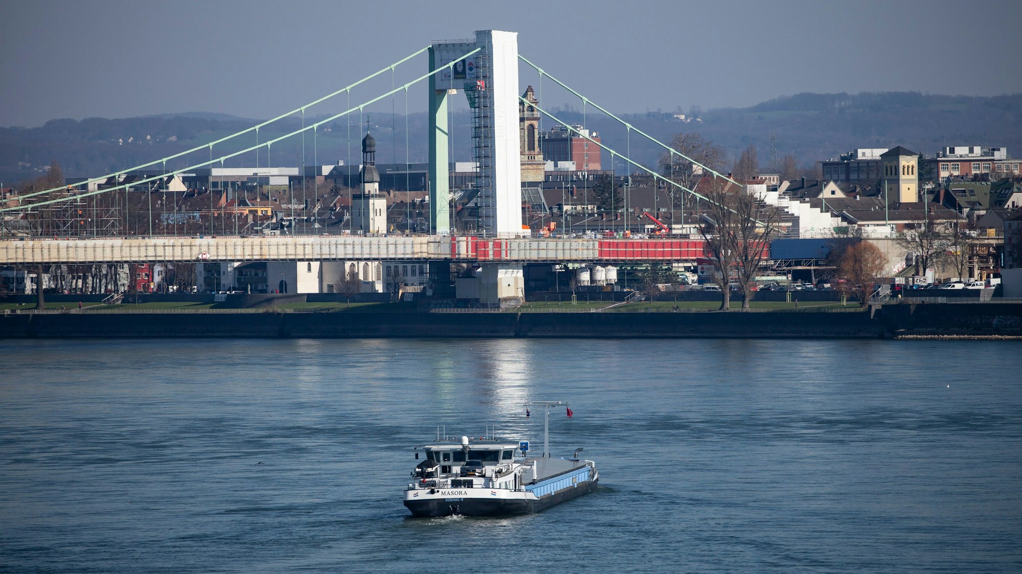 In der Entfernung ist die Brücke mit den Sanierungsmaßnahmen zu sehen. Im Vordergrund fährt ein Frachtschiff über den Rhein.