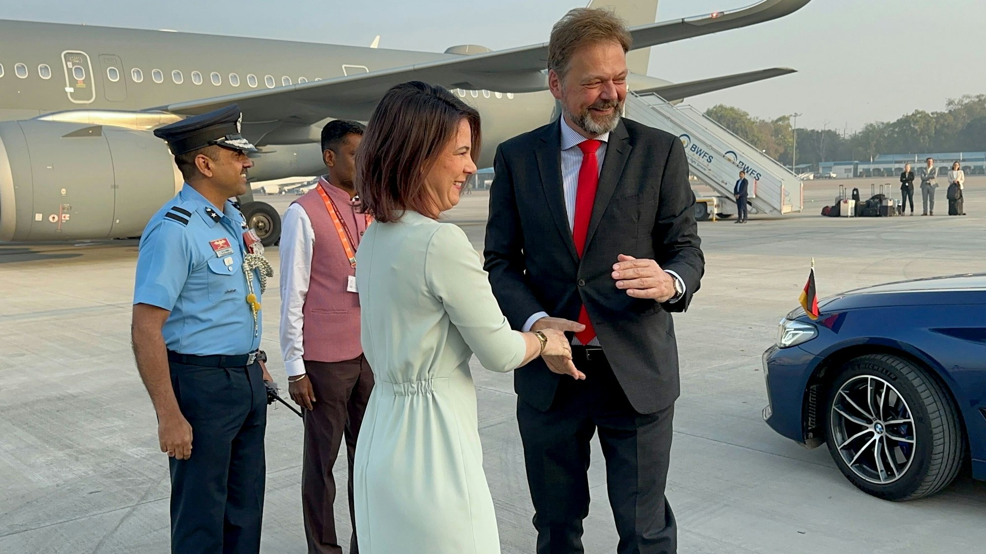 Annalena Baerbock und Philipp Ackermann stehen zusammen am Flughafen vor einem Flugzeug und geben sich die Hand.