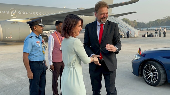 Annalena Baerbock und Philipp Ackermann stehen zusammen am Flughafen vor einem Flugzeug und geben sich die Hand.