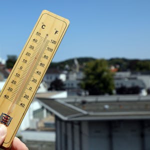 Jemand hält ein Thermometer in Bergisch Gladbach vor den strahlend blauen Himmel und die Häuserkulisse im Hintergrund.