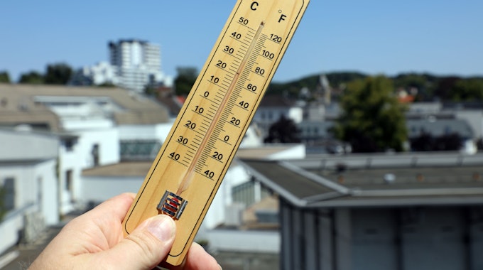 Jemand hält ein Thermometer in Bergisch Gladbach vor den strahlend blauen Himmel und die Häuserkulisse im Hintergrund.