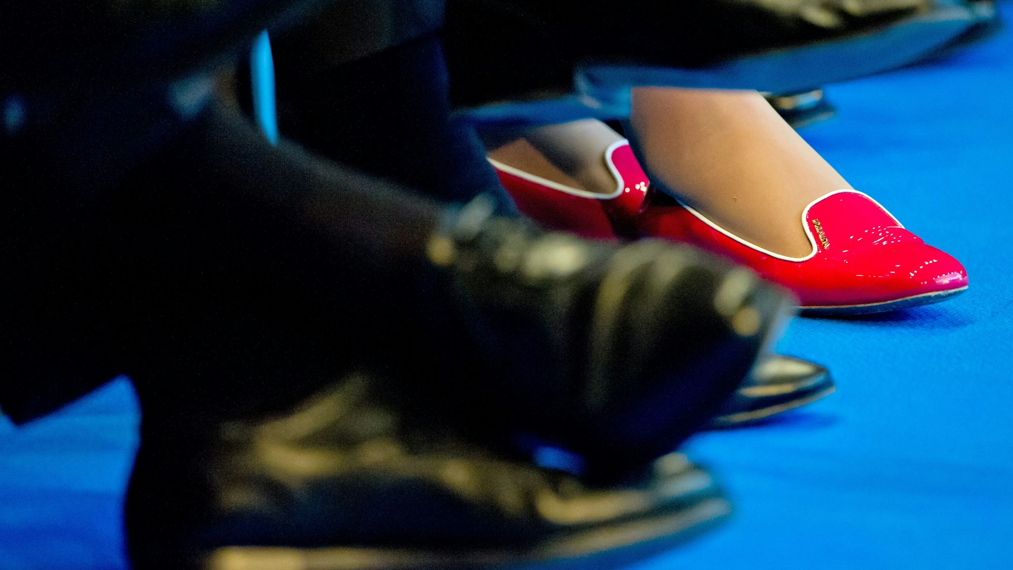 In roten Schuhen sitzt eine Teilnehmerin zwischen Männern in dunklen Anzügen und passendem Schuhwerk.