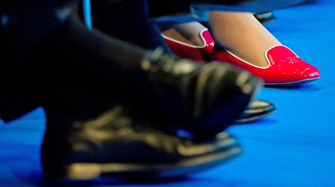 In roten Schuhen sitzt eine Teilnehmerin zwischen Männern in dunklen Anzügen und passendem Schuhwerk.
