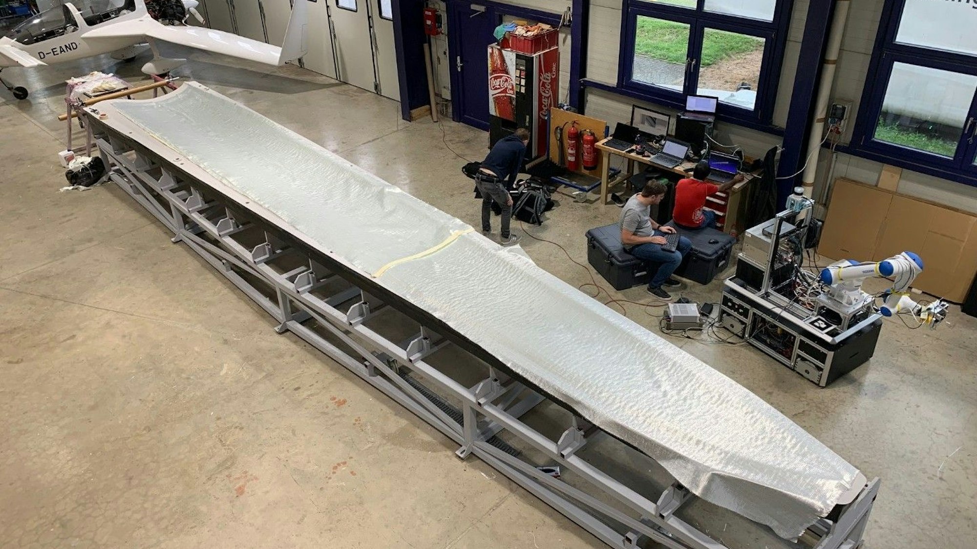 Bei der Aeroconcept GmbH ist für einen Test ein Faserpaket für eine Rotorblattspitze ausgelegt worden. Techniker sitzen neben der Form in einer Flugzeughalle.