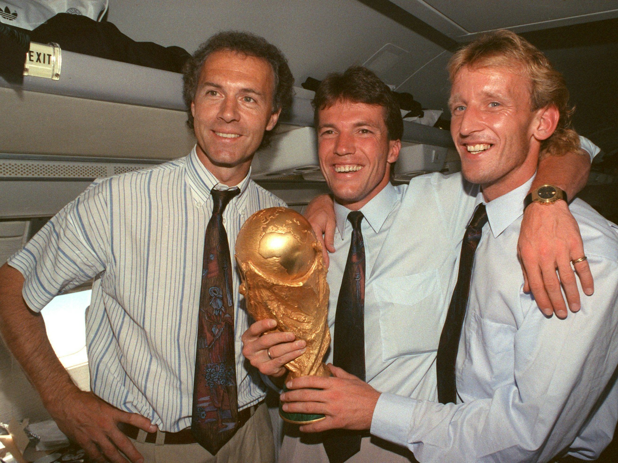 DFB-Teamchef Franz Beckenbauer, Kapitän und Mittelfeldspieler Lothar Matthäus und Abwehrspieler Andreas Brehme präsentieren strahlend den zwei Tage zuvor gewonnenen WM-Pokal im Flugzeug auf dem Rückflug von Rom nach Frankfurt.