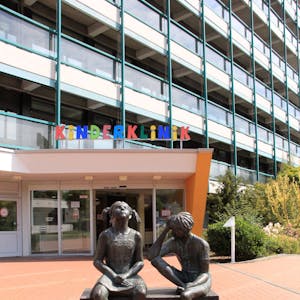 Über dem Eingang steht in bunten Buchstaben Kinderklinik. Im Vordergrund sind zwei Statuen von Kindern zu sehen.