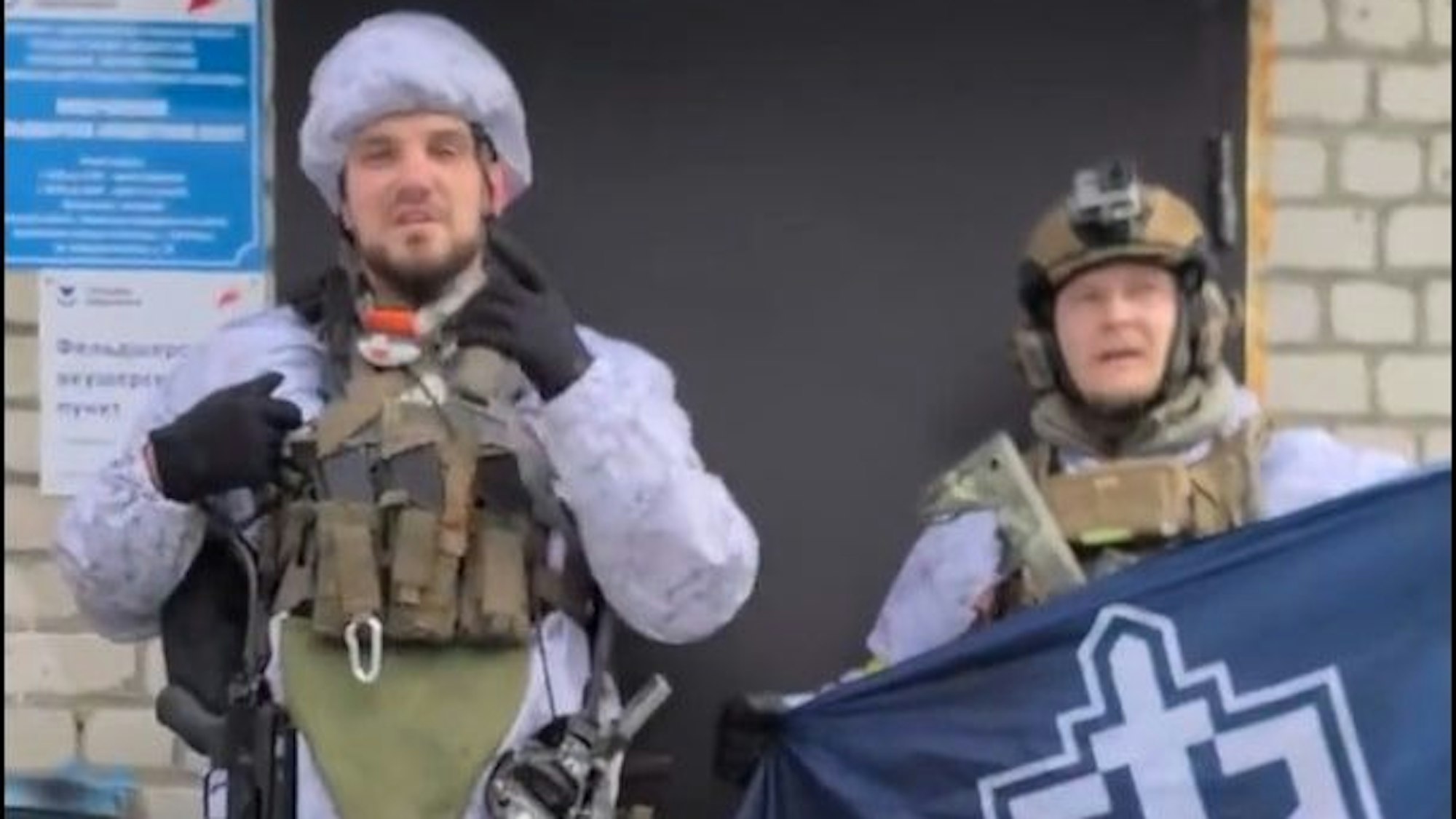 Ein etwas verpixelt Screenshot zeigt Denis Nikitin in Kampfmontur. Nikitin gestikuliert in Richtung Kamera. Neben ihm hält ein Mann - ebenfalls in Militärkleidung - ein blaues Banner mit dem Logo des Russischen Freiwilligenkorps hoch.