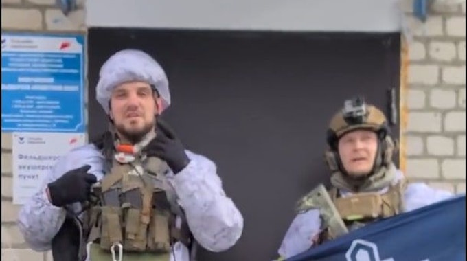 Ein etwas verpixelt Screenshot zeigt Denis Nikitin in Kampfmontur. Nikitin gestikuliert in Richtung Kamera. Neben ihm hält ein Mann - ebenfalls in Militärkleidung - ein blaues Banner mit dem Logo des Russischen Freiwilligenkorps hoch.&nbsp;