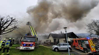 Über einem Haus steigt eine dichte, graue Rauchwolke auf. Von einer Drehleiter aus löschen Feuerwehrleute den brennenden Dachstuhl..