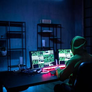 Ein Hacker mit Kapuze sitzt mit dem Rücken zur Kamera an einem Tisch. Das Licht ist ausgeschaltet, nur auf dem Bildschirm leuchtet es geheimnisvoll.