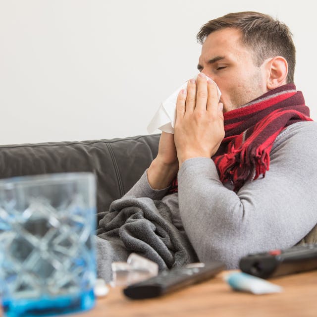 Ein Mann sitzt erkältet auf dem Sofa und putzt sich die Nase.