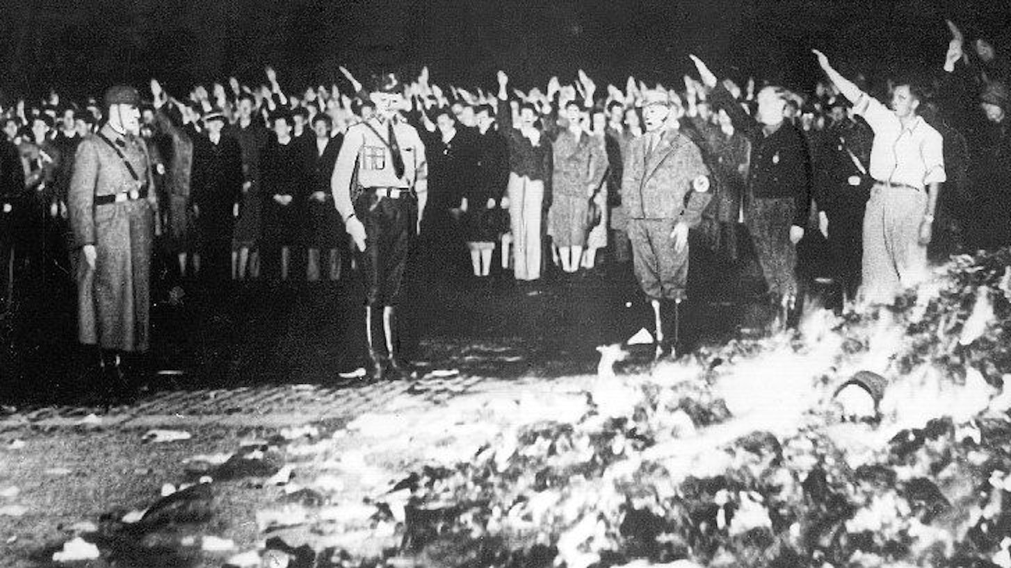 Das Archivbild zeigt eine Bücherverbrennung in Deutschland. Hinter einem brennenden Berg mit Büchern sind Menschen zu sehen, die den Hitlergruß zeigen.