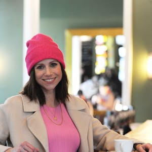Die Schauspielerin Katrin Heß sitzt am Tisch und trinkt Kaffee, sie trägt eine pinke Mütze und ein pinkes Kleid.