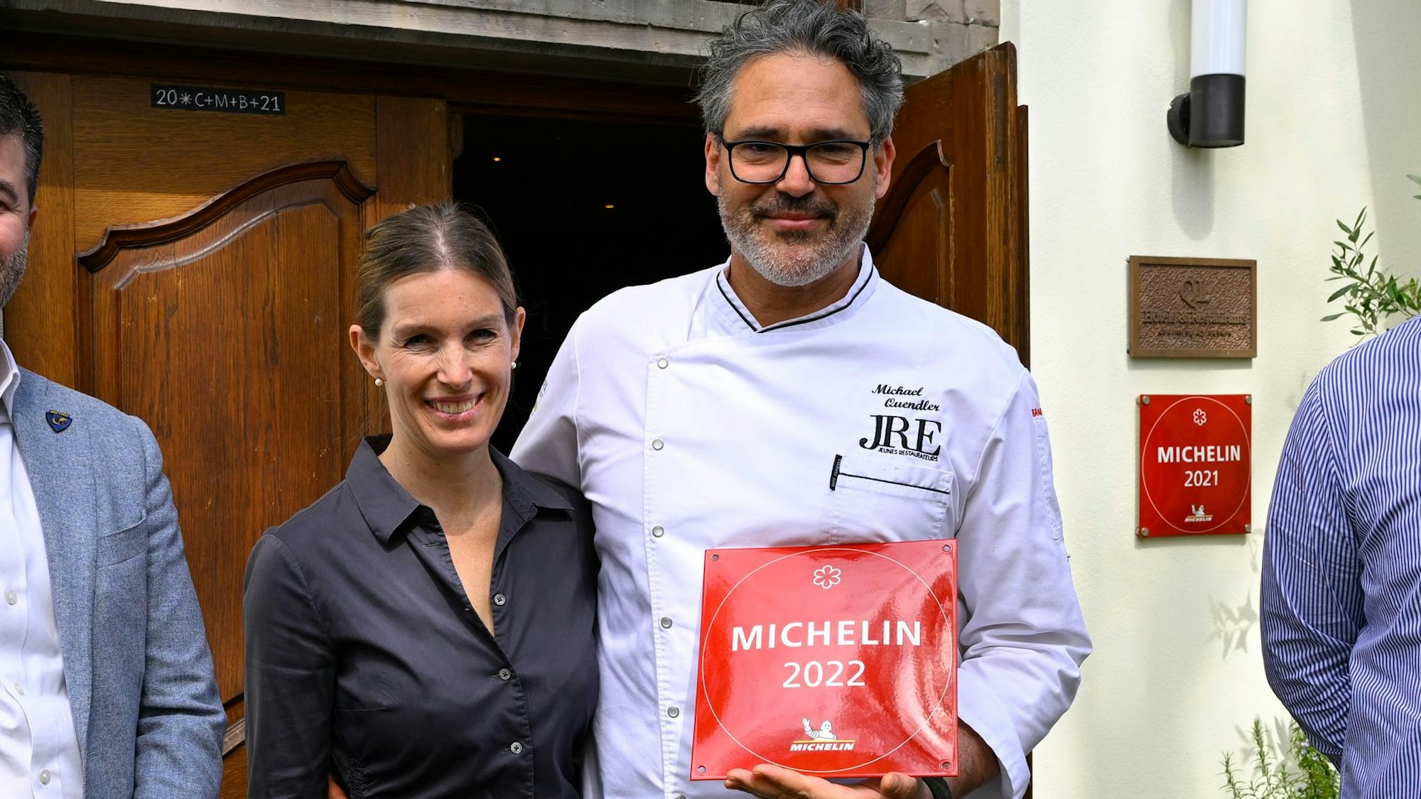 Michael und Birgitta Quendler stehen vor ihrem Restaurant und halten die Auszeichnung des Guide Michelin von 2022, eine rote Plakette.