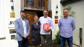 Vier Personen stehen vor einem Gebäude mit einer großen Holztüre. Ein Mann hält eine rote Plakette mit der Michelin-Auszeichnung in der Hand.
