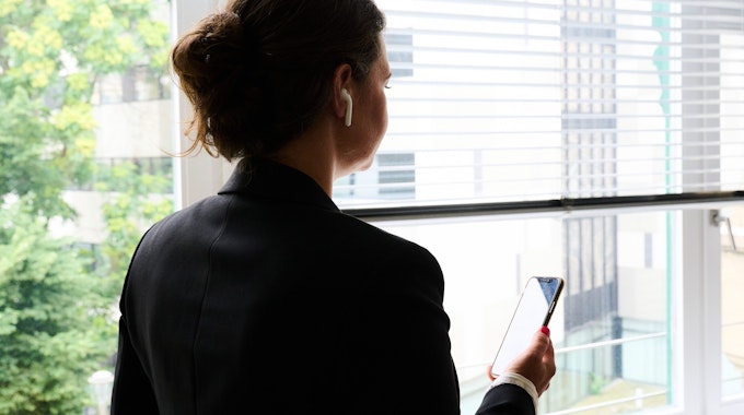 Eine Frau steht in einem Bürozimmer am Fenster und telefoniert über ein Headset. (gestellte Szene)&nbsp;