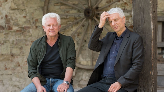 Die Schauspieler Miroslav Nemec und Udo Wachtveitl posieren für ein Foto. Beide Schauspieler sitzen auf einer Holzbank.