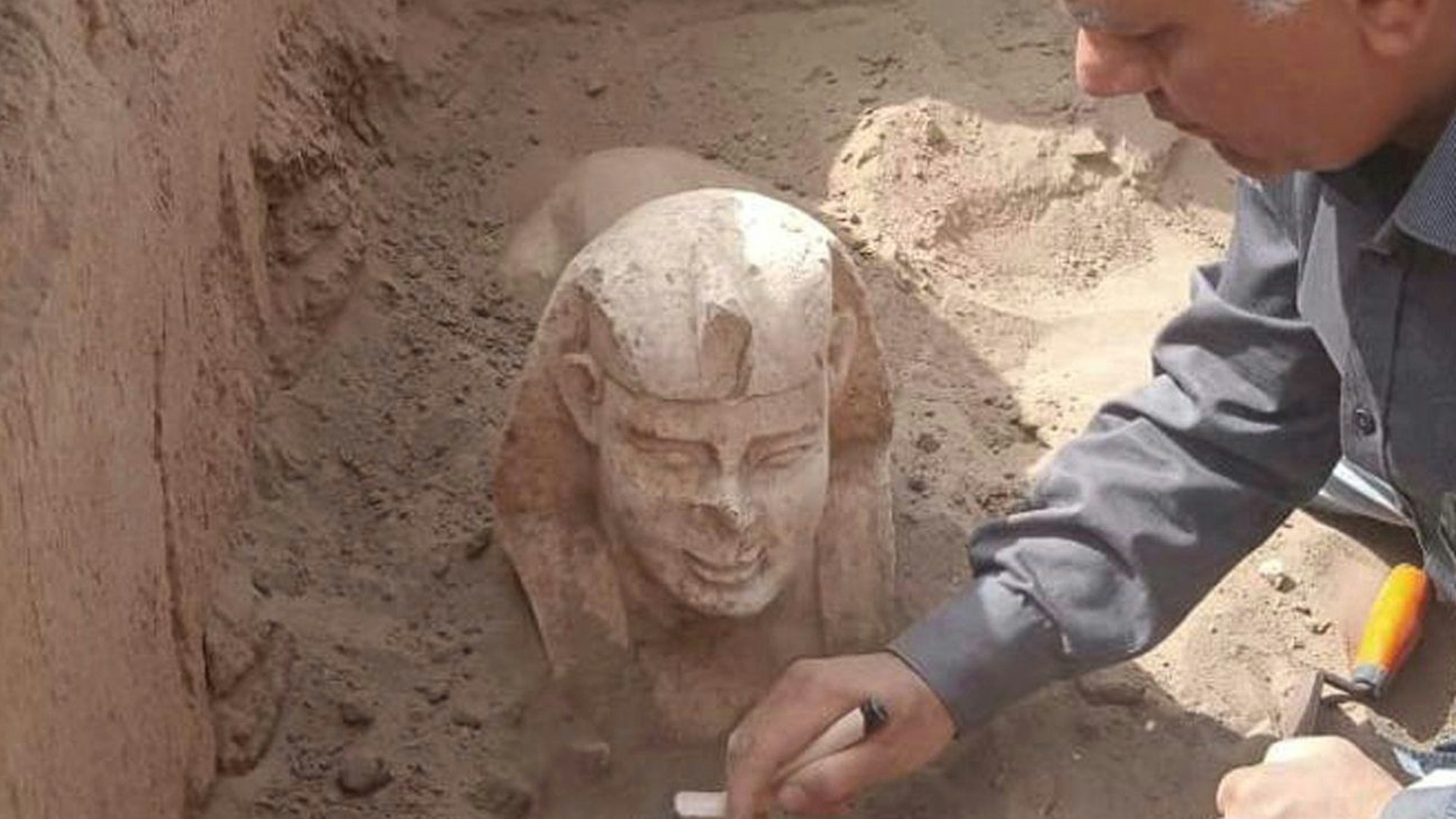Der Kopf einer Sphinx ragt aus dem Sand. Ein Archäologe beugt sich darüber.