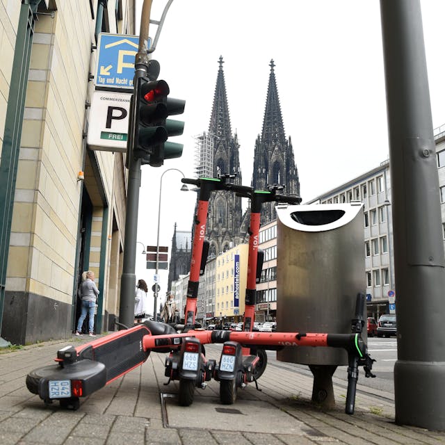 Drei E-Scooter stehen auf dem Bürgersteig. Im Hintergrund ist der Kölner Dom zu sehen.