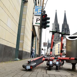 Drei E-Scooter stehen auf dem Bürgersteig. Im Hintergrund ist der Kölner Dom zu sehen.
