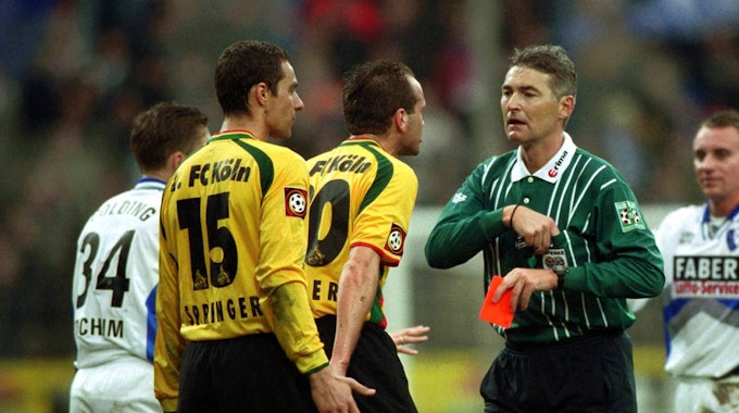 Zwei Spieler des 1. FC Köln in gelben Trikots diskutieren mit dem Schiedsrichter, der eine rote Karte in der Hand hält.