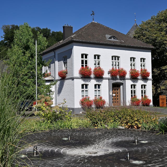 Das Rathaus in Odenthal mit roten Geranien vor den Fenstern. Vor dem Gebäude befindet sich ein Springbrunnen.
