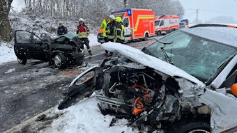 Ein völlig zerfetztes Auto liegt auf der Straßenseite im Schnee, quer auf der Straße steht ein anderer, ebenfalls schwer beschädigter Pkw. Mehrere Feuerwehrleute, Polizisten und Rettungsfahrzeuge stehen auf der winterlichen Straße.