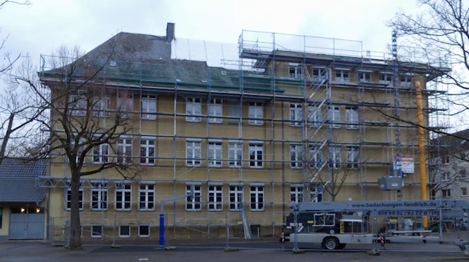 Ein mehrstöckiges großes Gebäude mit gelber Fassade ist zu sehen. Vor dem Haus stehen Gerüste, Teile des Dachs sind mit Planen zugedeckt.