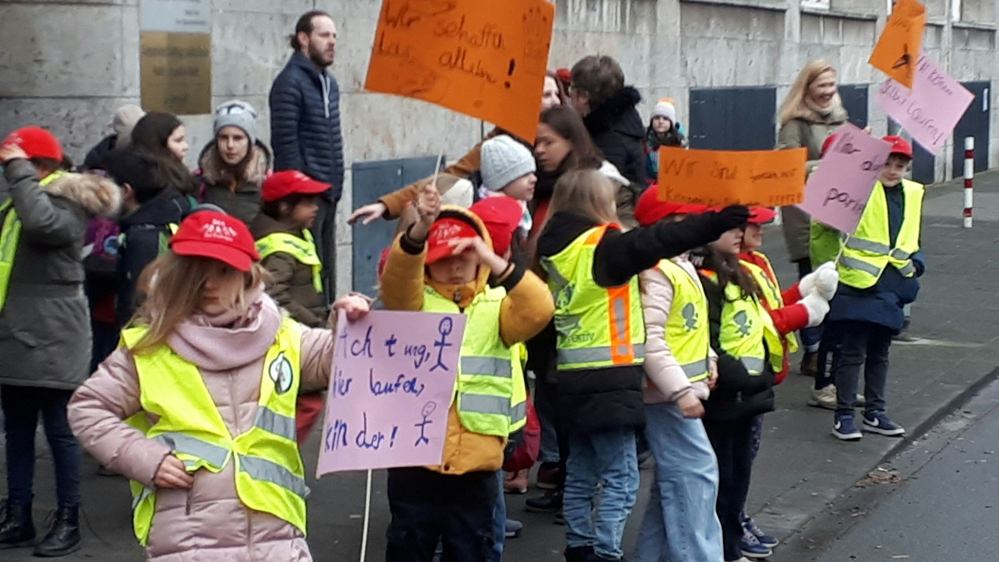Kinder mit Warnwesten, roten Kappen und Protestschildern vor dem Eingang eines Schulgebäudes