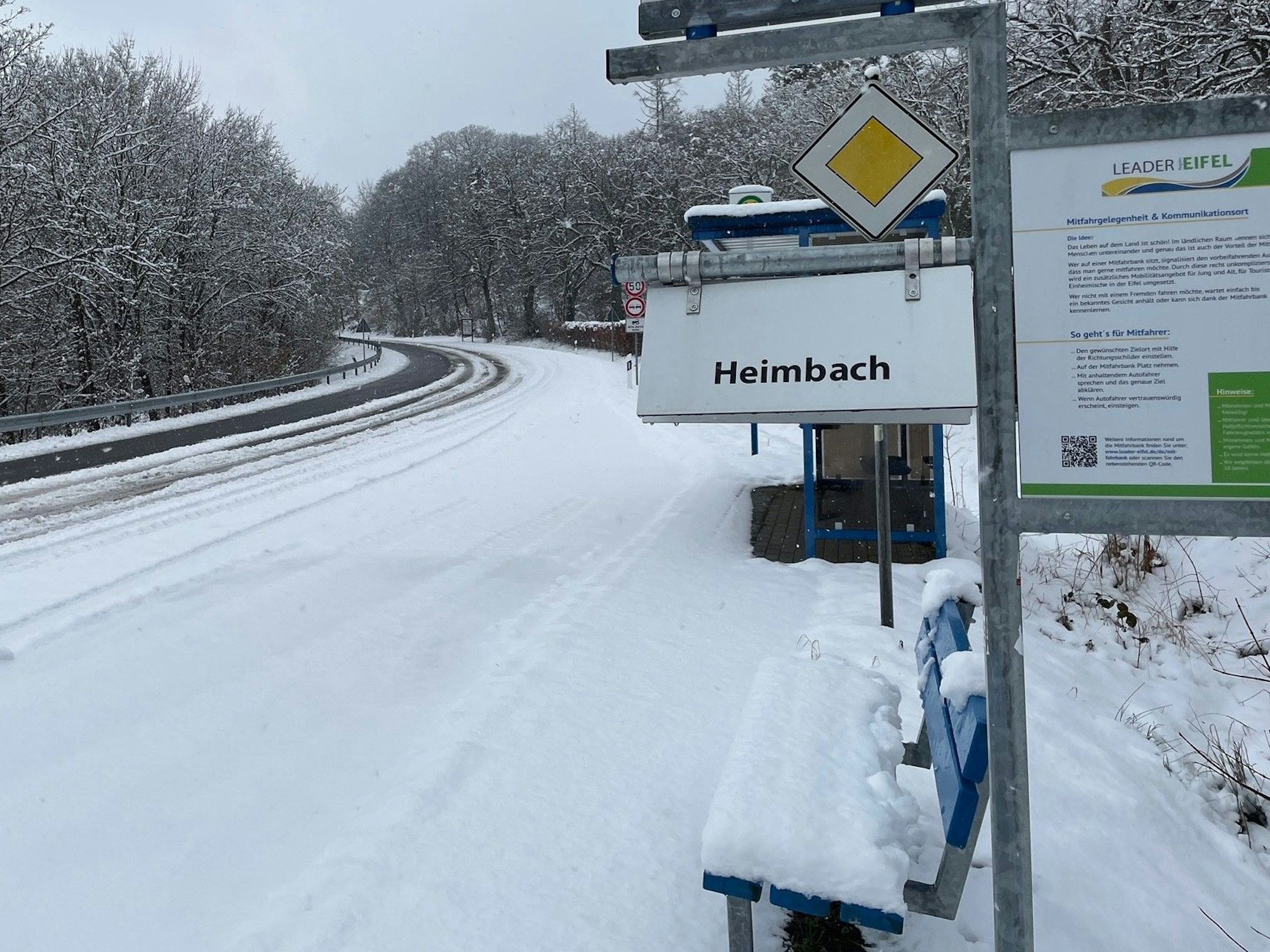 Eine Bank mit dem Hinweisschild "Heimbach" liegt unter einer Schneedecke.