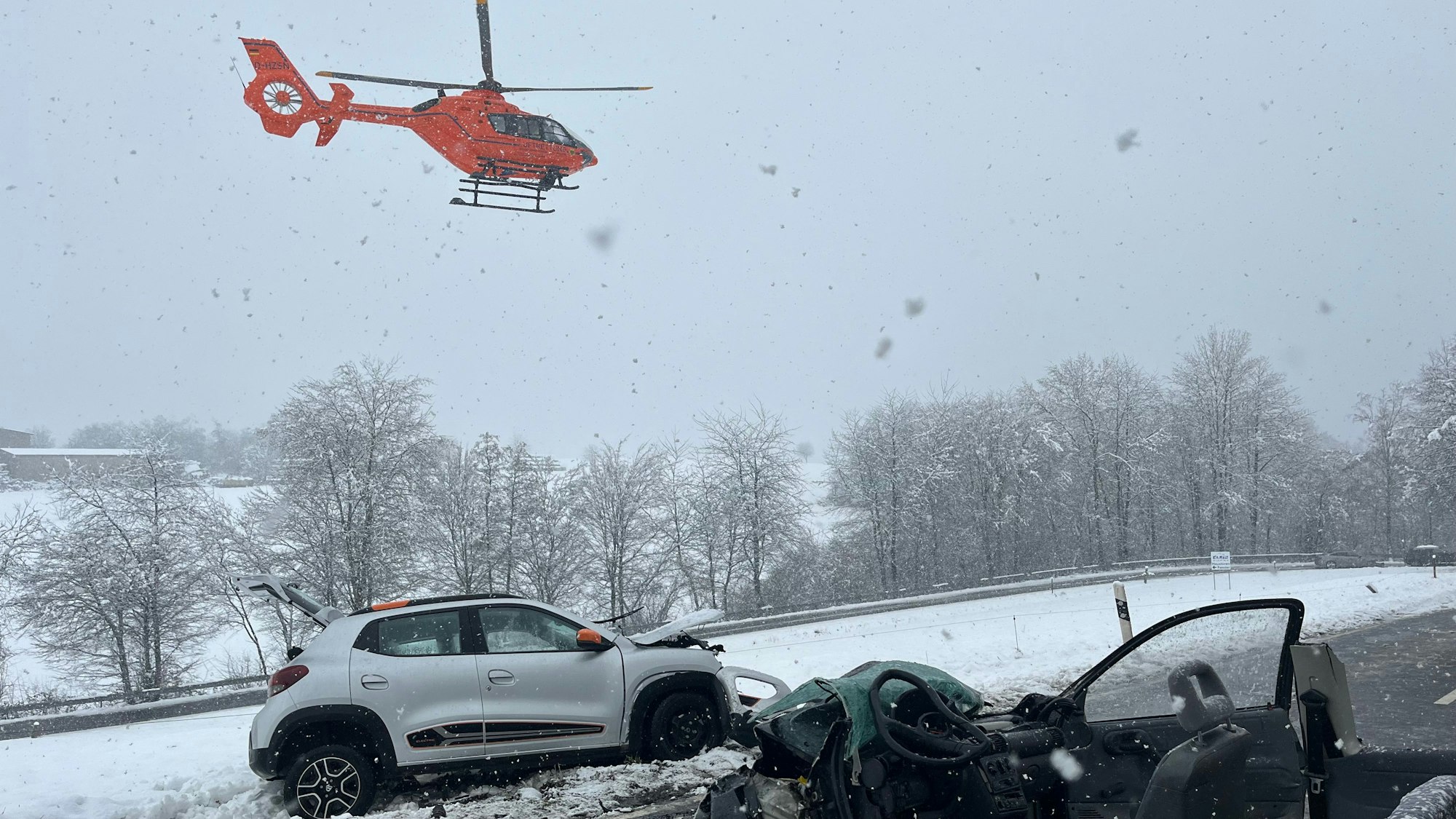 Ein Rettungshubschrauber fliegt über die verschneite Landschaft und die Straße, auf der die beiden Unfallfahrzeuge stehen.