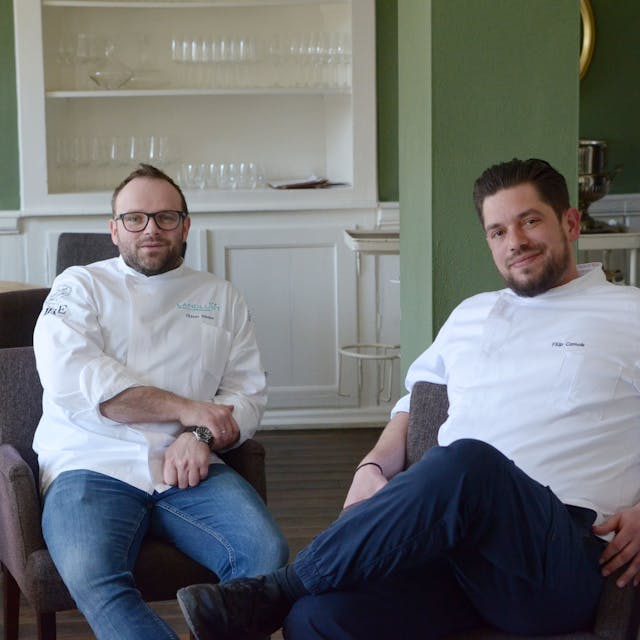 Der Geschäftsführer vom Bembergs Häuschen Oliver Röder und Küchenchef Filip Czmok sitzen in weißer Kochmontur im Gastraum ihres Restaurants.