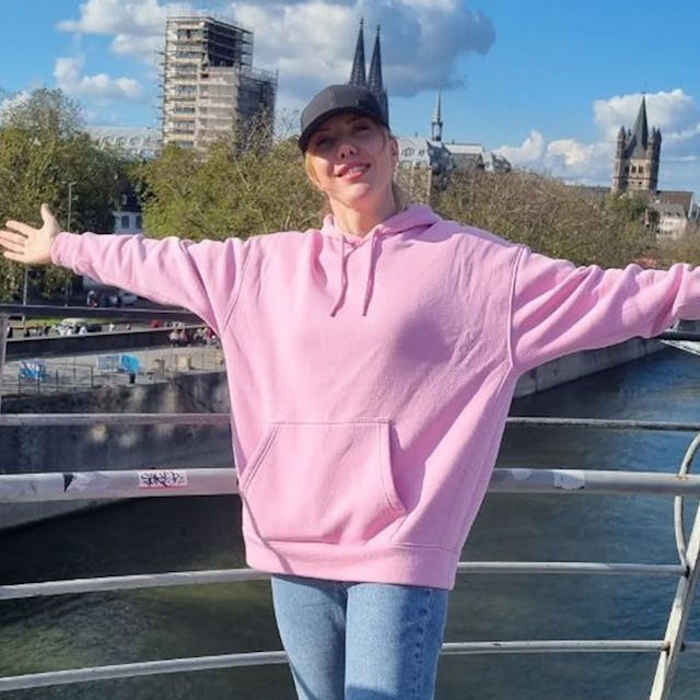 Eine Frau in einem rosa Sweater und ausgebreiteten Armen vor dem Rheinpanorama