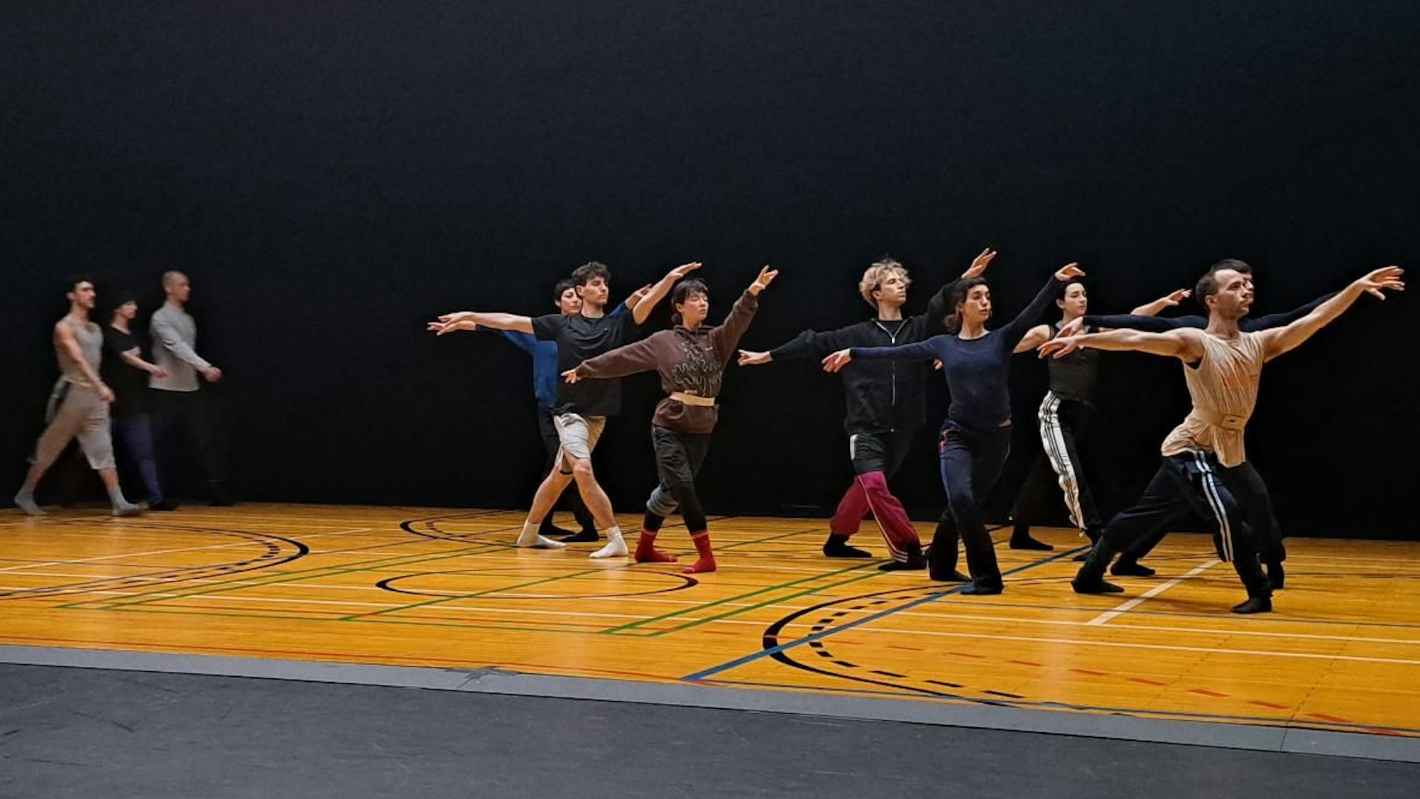 Eine Gruppe an Tänzerinnen und Tänzern steht in der gleichen Ballettposition in der Mitte der Bühne. Sie haben ausgestreckte Arme und Beine. Eine andere kleinere Gruppe geht im Hintergrund im Gleichschritt.