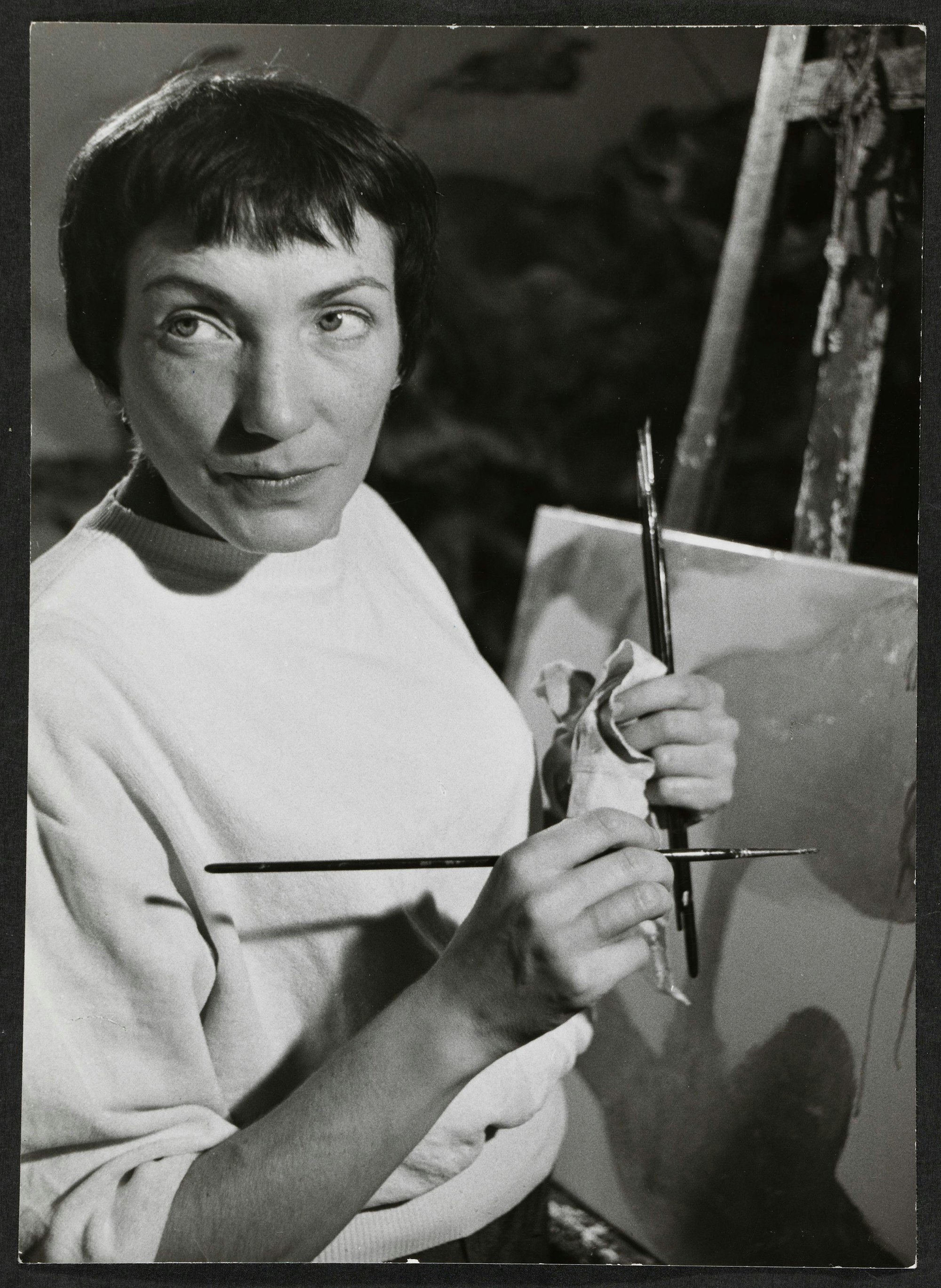 Die Künstlerin Ursula hält mehrere Pinsel in der Hand und steht vor einer Staffelei mit Bild.