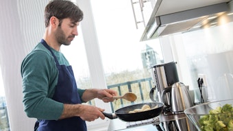 Ein Mann steht in einer Küche und schwenkt mit einem Kochlöffel in der Hand eine Pfanne.