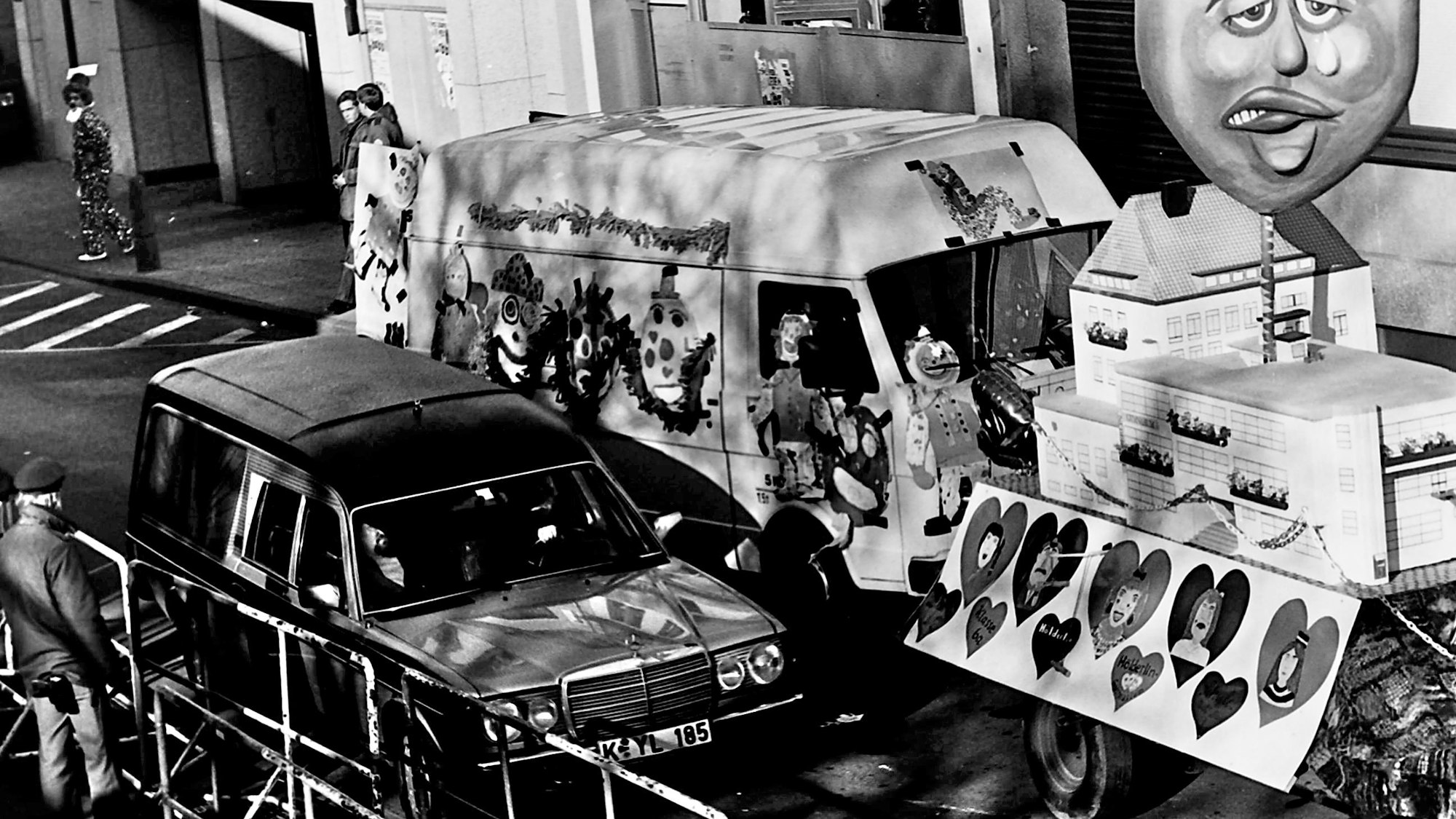Ein Leichenwagen fährt an einem Karnevalszug vorbei.