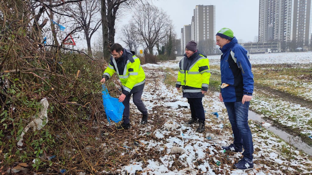 Hier pflücken drei städ­ti­sche Mit­ar­bei­ter Plas­tik­müll aus den Sträu­chern in Köln-Me­sche­nich.