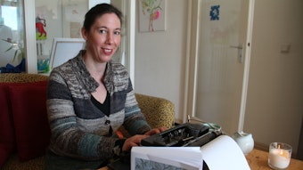 Nießen sitzt an einer Schreibmaschine mit ihrem Buch daneben und blickt in die Kamera.