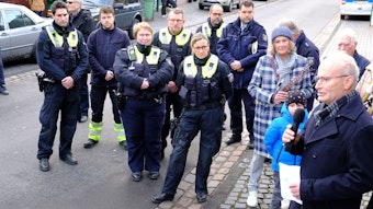 Polizistinnen und Polizisten sowie Menschen in ziviler Kleidung stehen auf einer Straße und hören Michael Heinen zu.