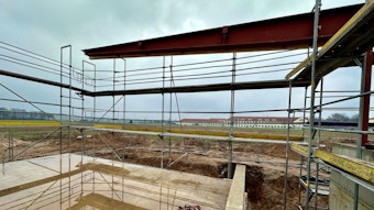 Ackerfläche mit aufgestelltem Gerüst. Flaches Altbau-Schulgebäude im Hintergrund