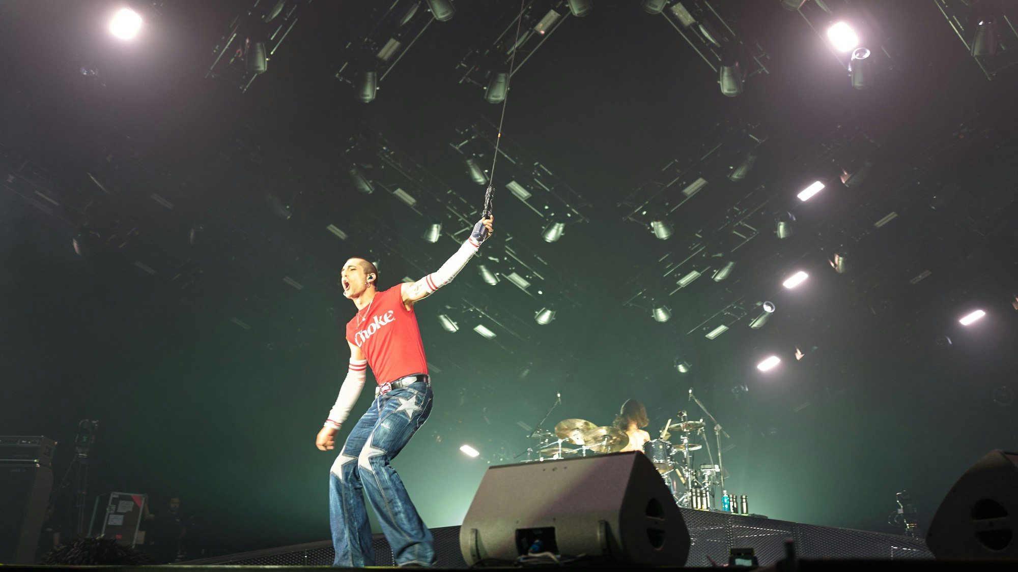 Måneskin-Frontman Damiano David steht auf der Bühne der Lanxess-Arena. Er hält das von der Decke hängende Mikrofon und schreit ins Publikum. Hinter im ist Schlagzeiger Ethan Torchio.
