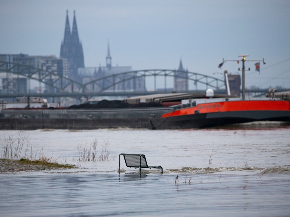 Binnenschiffe fahren auf dem Rhein vor der Kulisse des Kölner Doms, im Vordergrund steht eine vom Wasser umspülte Sitzbank. Noch hat der Kölner Pegel die Hochwassermarke nicht überschritten.