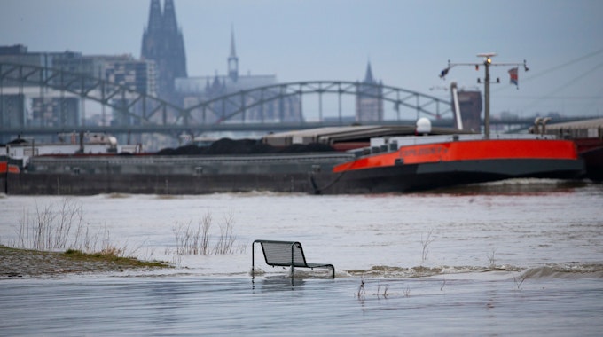 Binnenschiffe fahren auf dem Rhein vor der Kulisse des Kölner Doms, im Vordergrund steht eine vom Wasser umspülte Sitzbank. Noch hat der Kölner Pegel die Hochwassermarke nicht überschritten.