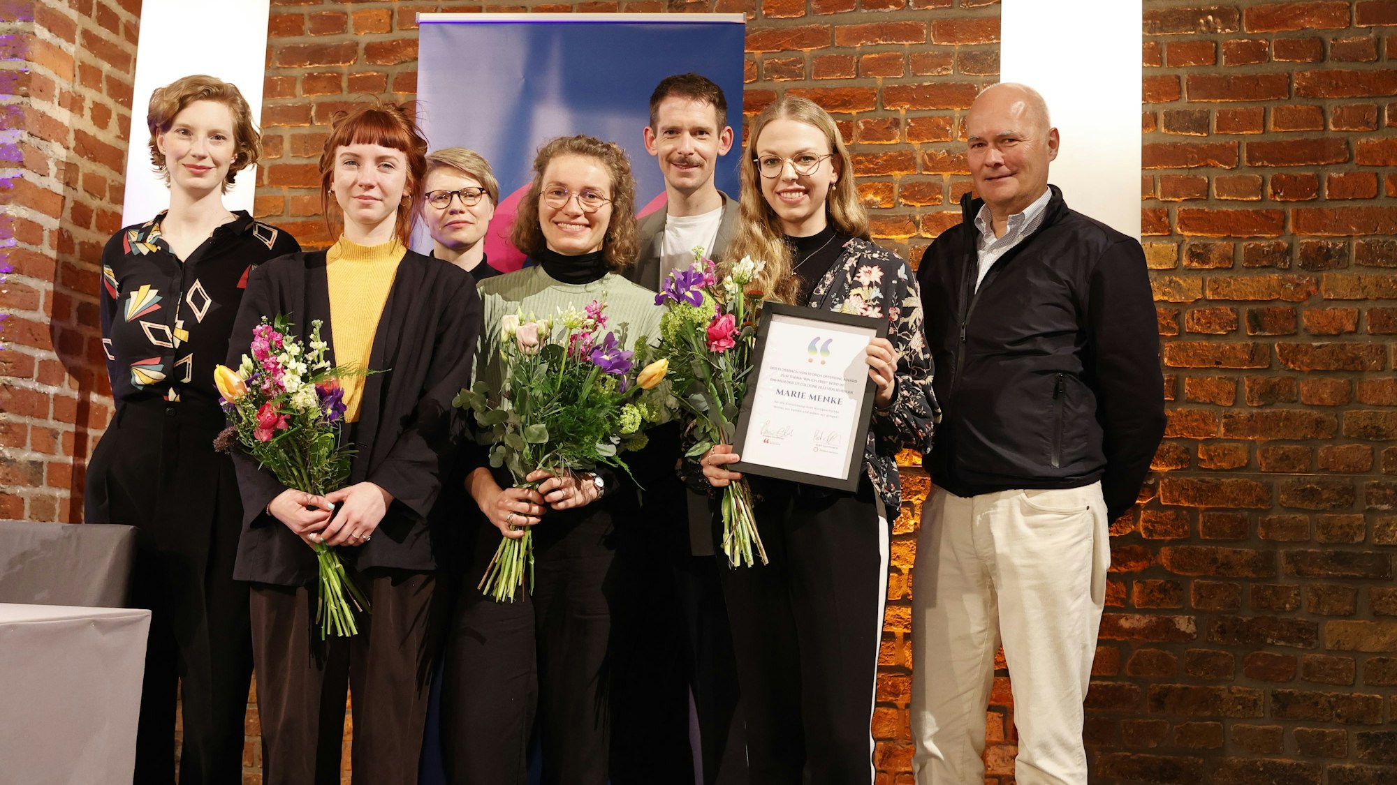 Luise Wolfram (v. links), Leonie Ziem, Nadja Küchenmeister, Verena Schieber, Benni Bauerdick, Marie Menke und Kurt von Storch lächeln in die Kamera. Die Finalistinnen haben Blumensträuse in der Hand, Gewinnerin Marie Menke dazu eine Urkunde.