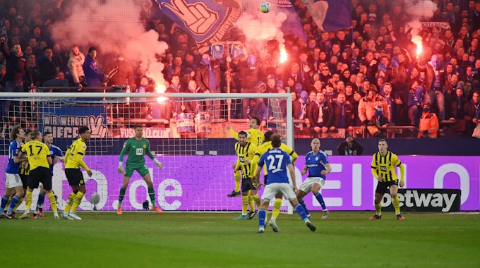 Spieler von Schalke und Dortmund kämpfen um den Ball, während im Hintergrund Fans bengalische Feuer abbrennen.