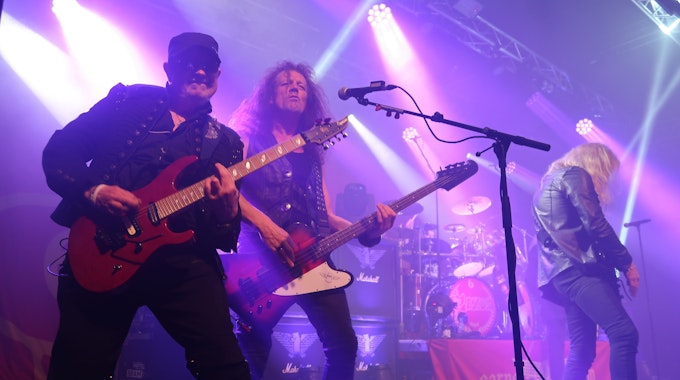 Ein Gitarrist und ein Musiker posieren auf einer in violettes Licht getauchten Bühne für den Fotografen.