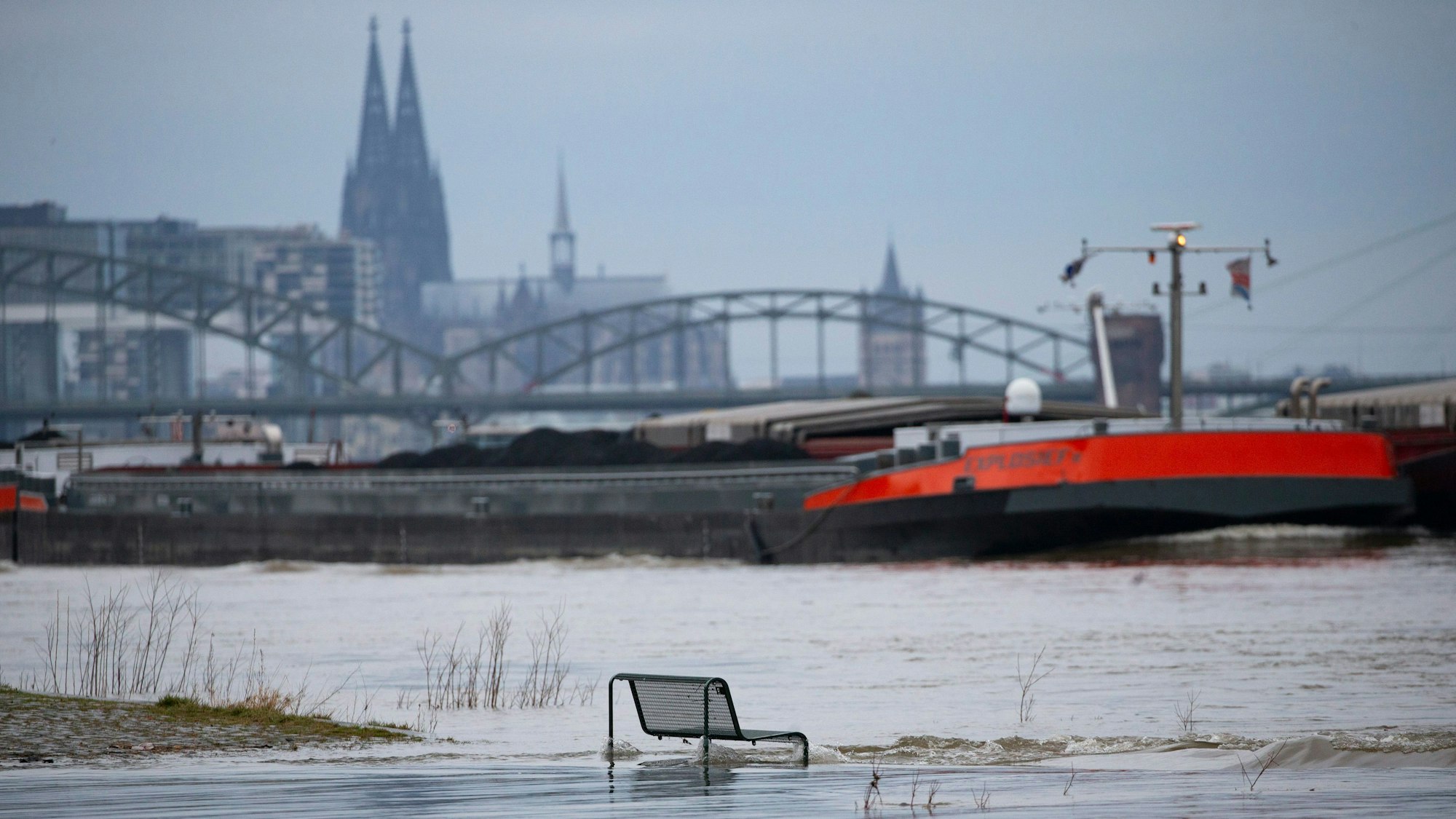 Binnenschiffe fahren auf dem Rhein vor der Kulisse des Kölner Doms, im Vordergrund steht eine vom Wasser umspülte Sitzbank.